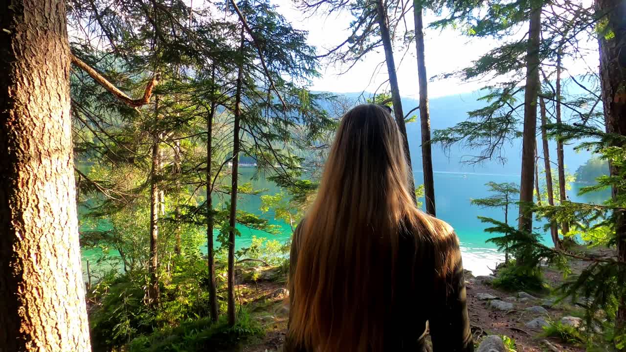 Third person view of a blonde girl in a green dress, walking through a forest until she reaches the shore of the famous lake Eibsee in Germany
