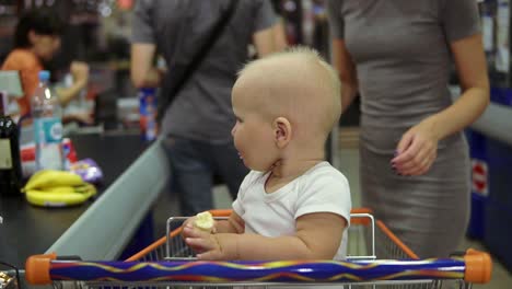 Portrait-of-a-cute-little-baby-taking-banana-and-sitting-in-a-shopping-cart-while-her-mother-is-taking-out-products-from-the