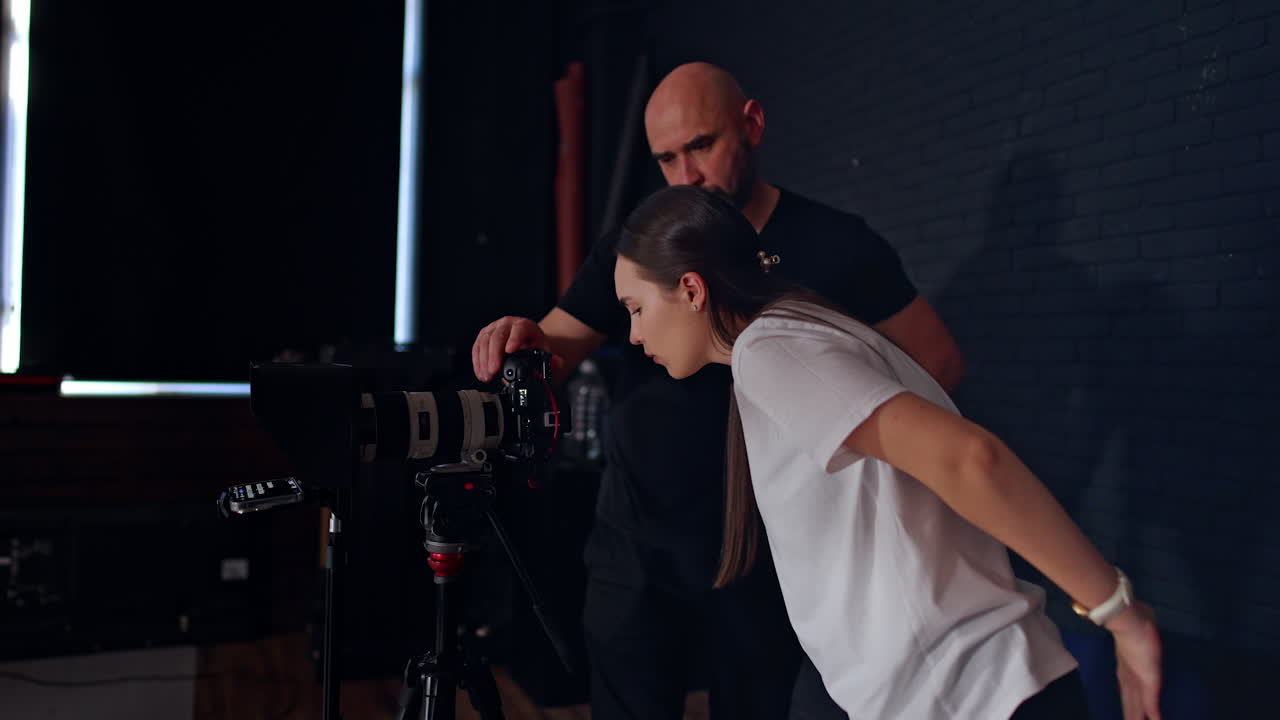 Long-haired Caucasian woman in white t-shirt leans closer to camera display and prompter. Bearded photographer in black t-shirt stands beside. Photo studio backstage.