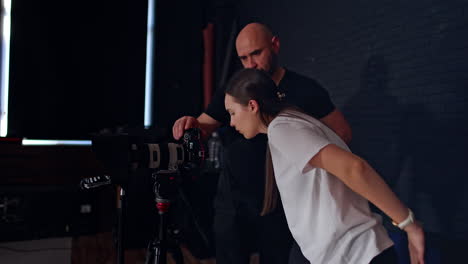 long-haired caucasian woman in white t-shirt leans closer to camera display and prompter. bearded photographer in black t-shirt stands beside. photo studio backstage.