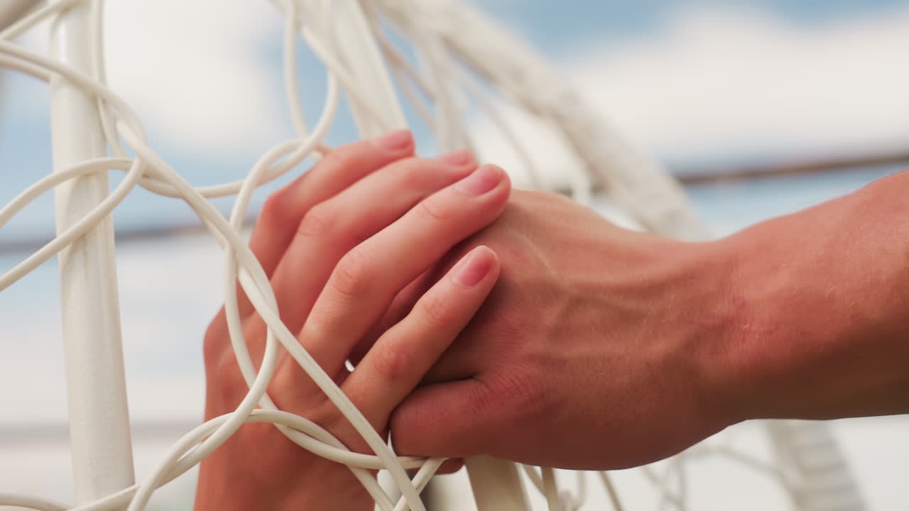 romantic couple hands entwined through net sunlit closeup of two hands clasped amid hammock rope against soft blue ocean and sky intimate supportive gesture by vacationing couple on beachside