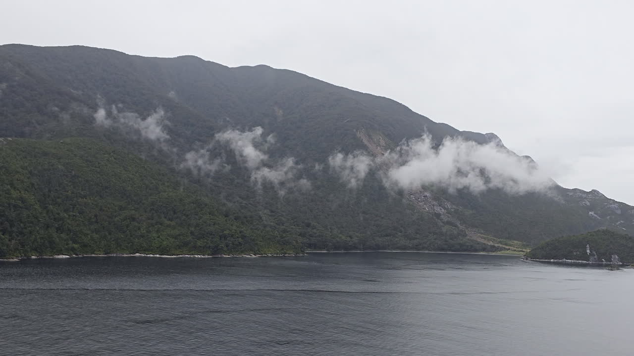 Milford Sound vista of steep, forested mountains shrouded in low clouds and mist, dark calm water reflecting the moody, remote fiord landscape in Fiordland National Park