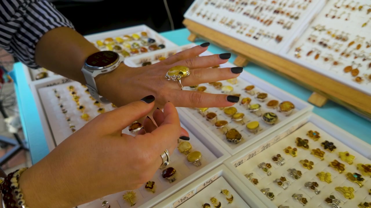 Primer plano de las manos de una mujer examinando y probándose anillos y joyas de ámbar auténtico en un puesto de mercado tradicional en Gdynia