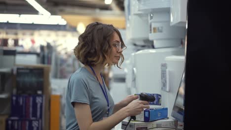 Curly-haired-slender-woman-seller-consultant-in-household-department-scan-by-barcode-stationery-goods-in-the-boxes.-Side-view