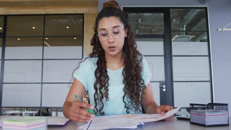 Happy-biracial-creative-businesswoman-at-desk,-reading-notes-and-talking-during-video-call