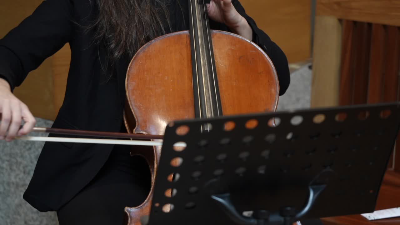 Person playing cello with hands on strings and fingerboard while reading sheet music in indoor performance setting
