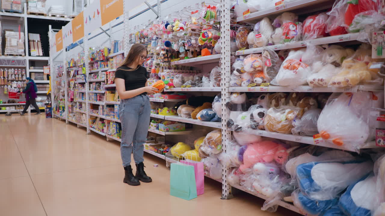 mujer con camiseta negra y vaqueros comprando en una tienda de juguetes, alcanzando un juguete de peluche suave en el estante, coloridos animales de relleno y juegos se alinean en los pasillos mientras las bolsas de compras pastel descansan en el suelo