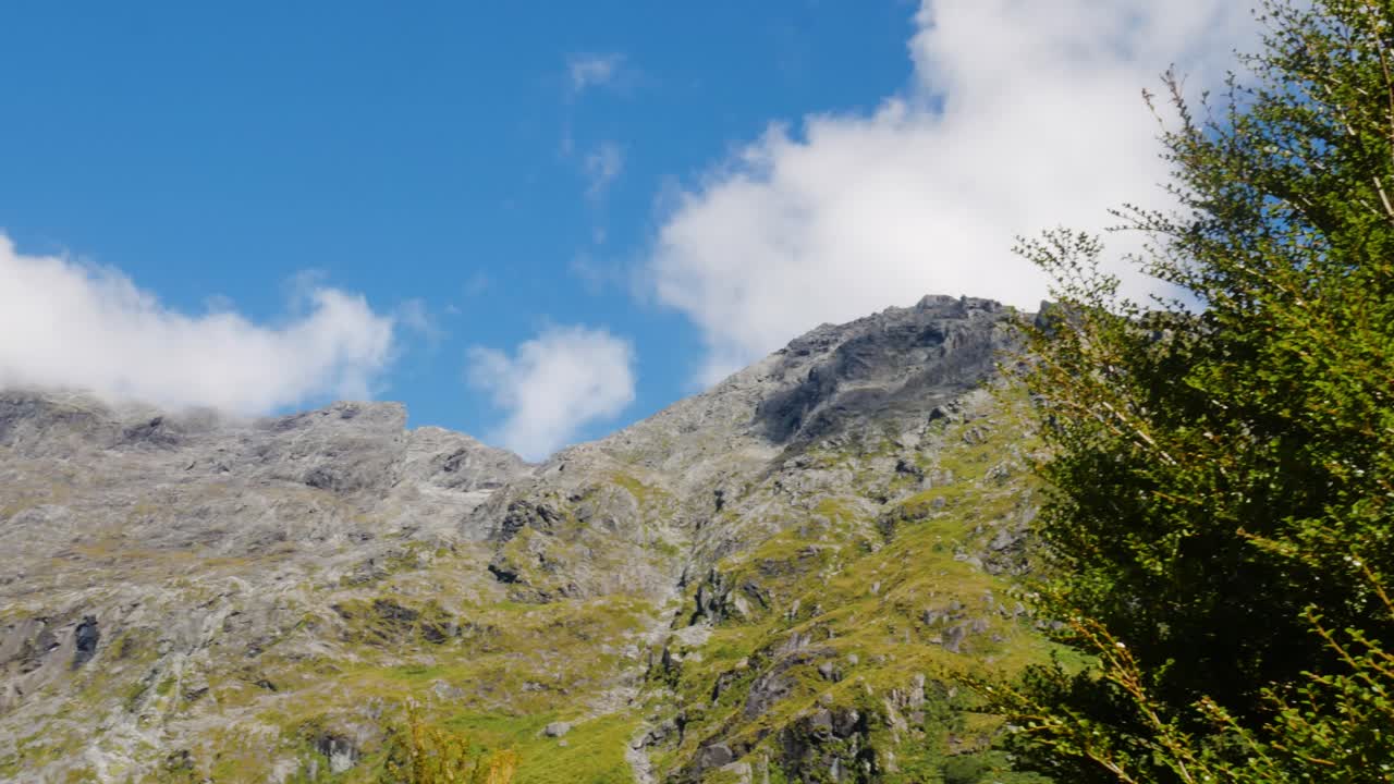 Bottom up shot of hovering white clouds over high mountain peaks during sunny day and blue sky - Getrude Saddle Path,New Zealand
