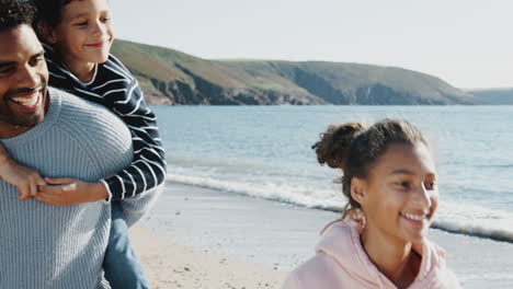 family walking along autumn beach together with father giving son piggyback ride
