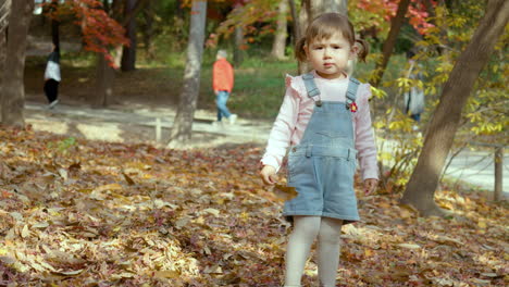 a cute two years old girl wearing denim jumper holding an autumn leaf looking at the camera at the parl