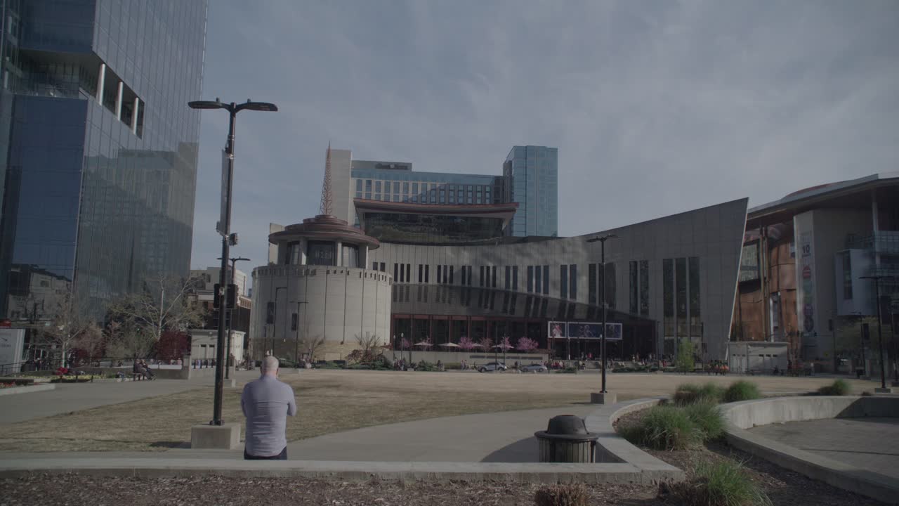 A wide shot of an urban park with contemporary buildings, including a distinctive one with an angular design, against a blue sky with sparse clouds, with a solitary figure seated