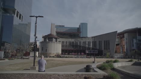 A-wide-shot-of-an-urban-park-with-contemporary-buildings,-including-a-distinctive-one-with-an-angular-design,-against-a-blue-sky-with-sparse-clouds,-with-a-solitary-figure-seated