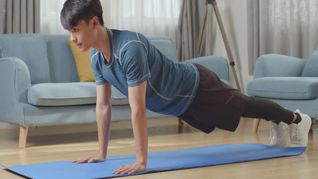 운동복을 입은 근육질, 체력 좋은 아시아인 남성이 집에서 활기차게 밀어붙이는 운동을 하고 있다.