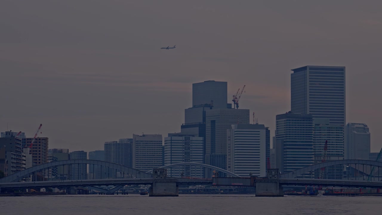 Un avión de pasajeros cruza sobre el denso horizonte comercial de Tokio al anochecer, con el Puente Kachidoki arqueándose sobre el oscuro río Sumida debajo, y grúas de construcción y torres de oficinas en un sombrío gris-púrpura.