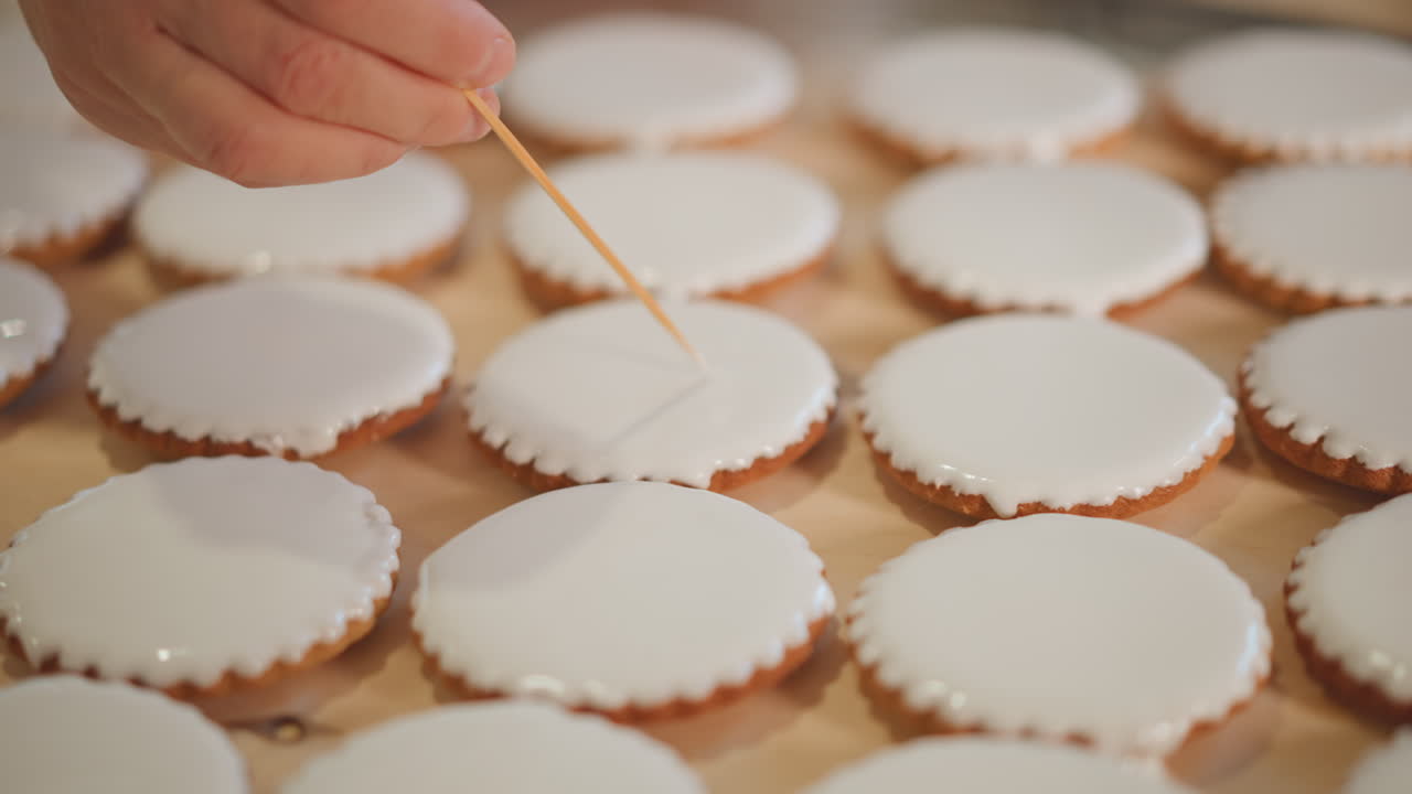 Rows iced mini pies being decorated by quick skilled hands, repetitive pattern across wooden trays, highvolume bakery setting, rhythmic motion, careful dotting of glaze, warm neutral lighting
