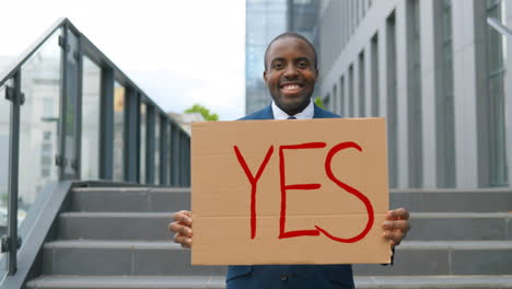 portrait of african american elegant young man in blue suit smiling and showing yes" signboard in the street"