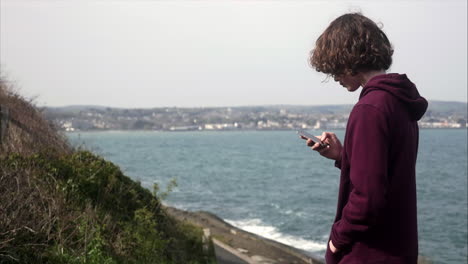 joven adolescente buscando en las redes sociales en su teléfono móvil con vista costera en segundo plano, copiando espacio