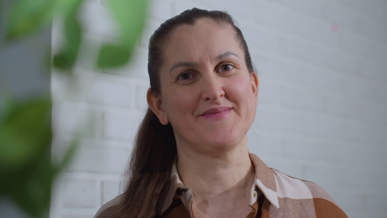 Nature enthusiast smiles while touching hair, standing in front of white brick wall with blurred green plant at side, warm indoor lighting highlighting relaxed expression and casual plaid shirt style