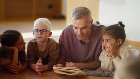 A-male-teacher-with-gray-hair-in-a-purple-T-shirt-reads-a-book-to-preschool-children-who-lie-on-the-floor-on-special-pillows-in-a-club-for-preparing-children-for-school-in-a-cozy-room