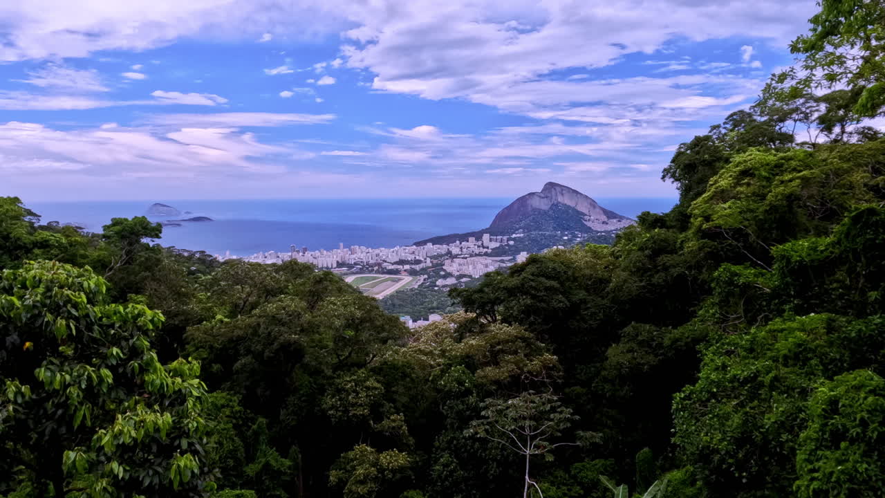 Aerial time-lapse overlooking Rio's equestrian center nestled in lush forest. Features the city skyline, ocean, and Two Brothers Hill under a dynamic blue sky with moving clouds
