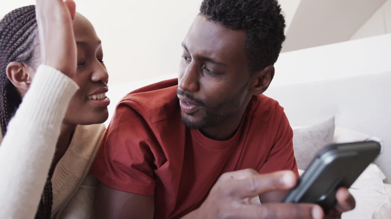 una feliz pareja afroamericana acostada en la cama usando un teléfono inteligente y hablando en casa, en cámara lenta