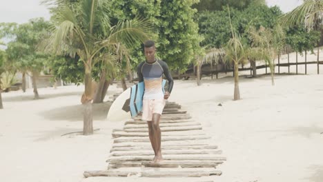 happy african american man carrying surfboard running on boardwalk on sunny beach, slow motion