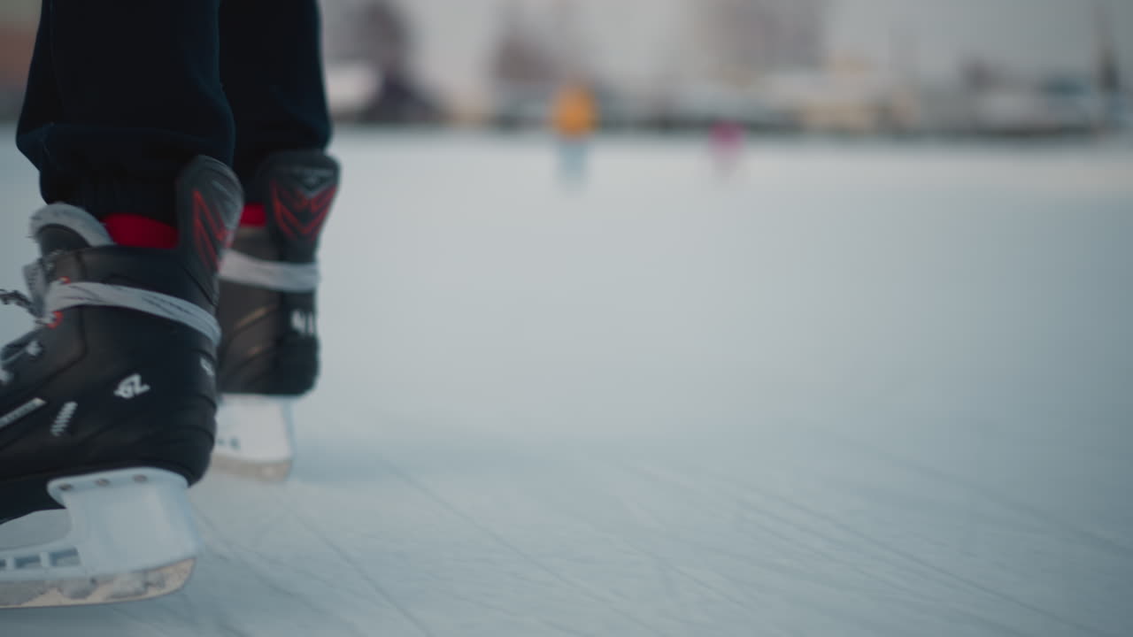 close up of skillful skater boots gliding on ice rink with blurred people having fun near residential buildings under pale winter light, ice surface marked by blade scratches and soft snow banks