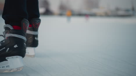 close up of skillful skater boots gliding on ice rink with blurred people having fun near residential buildings under pale winter light, ice surface marked by blade scratches and soft snow banks