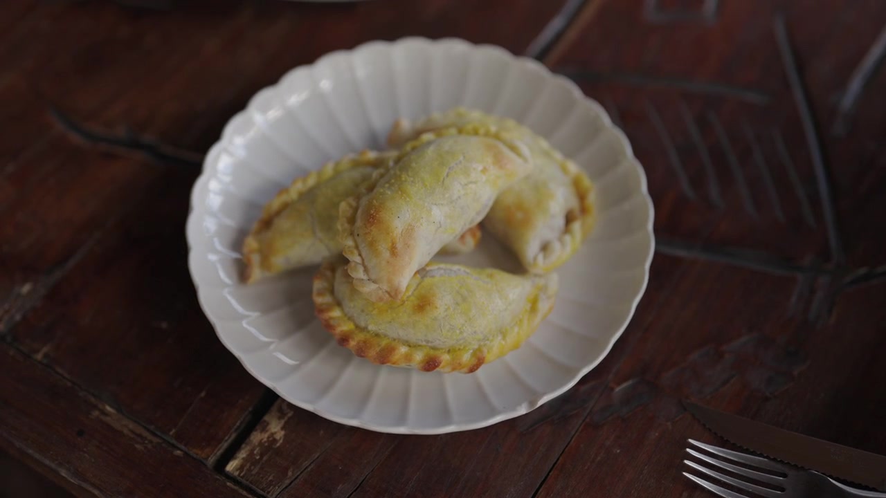 Tre empanadas dorate sono adagiate su un piatto bianco sopra un tavolo di legno. Ideale per blog culinari o approfondimenti sulla cucina culturale