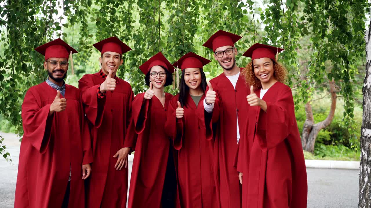 Portrait of excited graduating students multiethnic group standing outdoors in red gowns and mortar-boards and talking then showing thumbs-up and looking at camera.