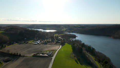 Aerial-shot-showing-agricultural-fields-beside-blue-lake-and-bright-sunset-in-backdrop