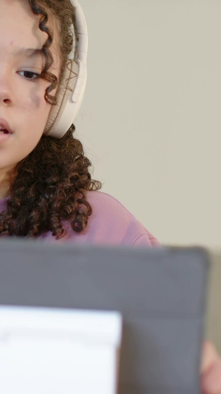 Vertical video: Reading woman with headphones and pink pullover at desk, using tablet, copy space