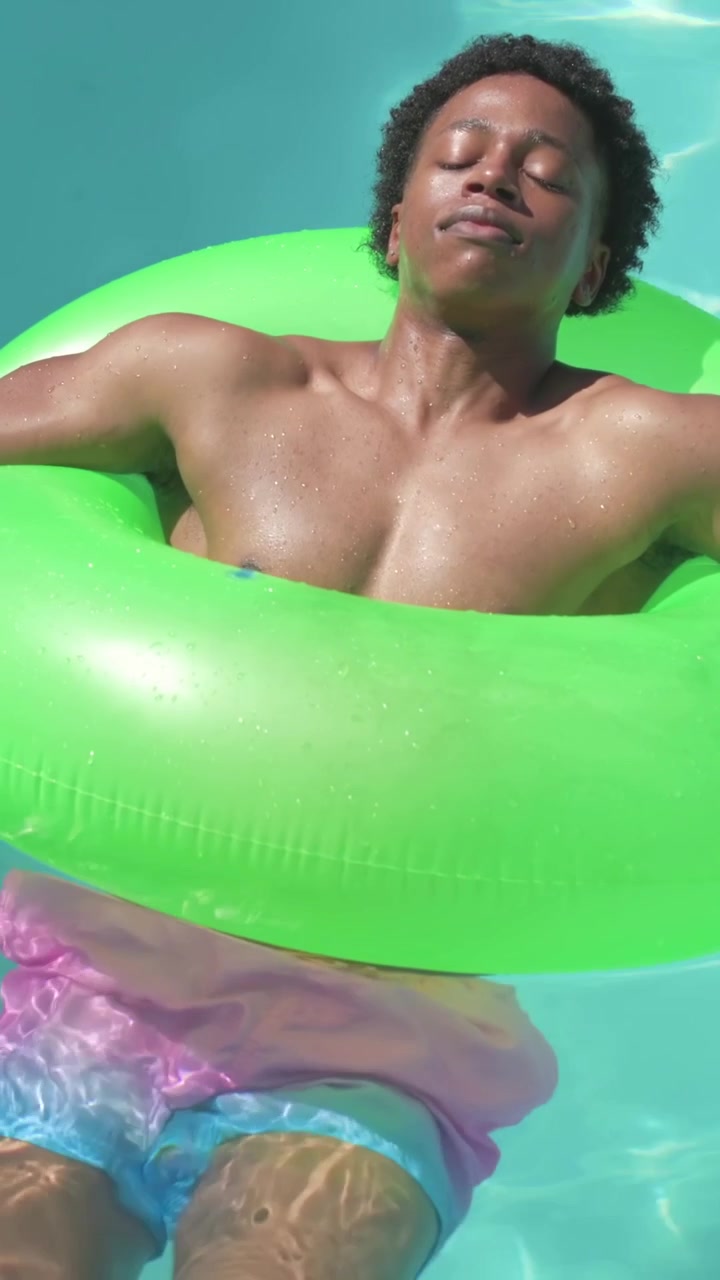 Vertical video: Camera panning man reclining in pool on green tube, soaking up sun in pastel trunks