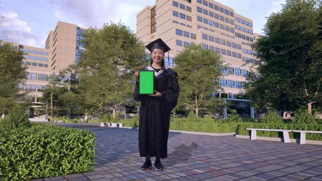 full body of asian woman student graduates in cap and gown smiling and showing green screen tablet to the camera in front of a magnificent university building