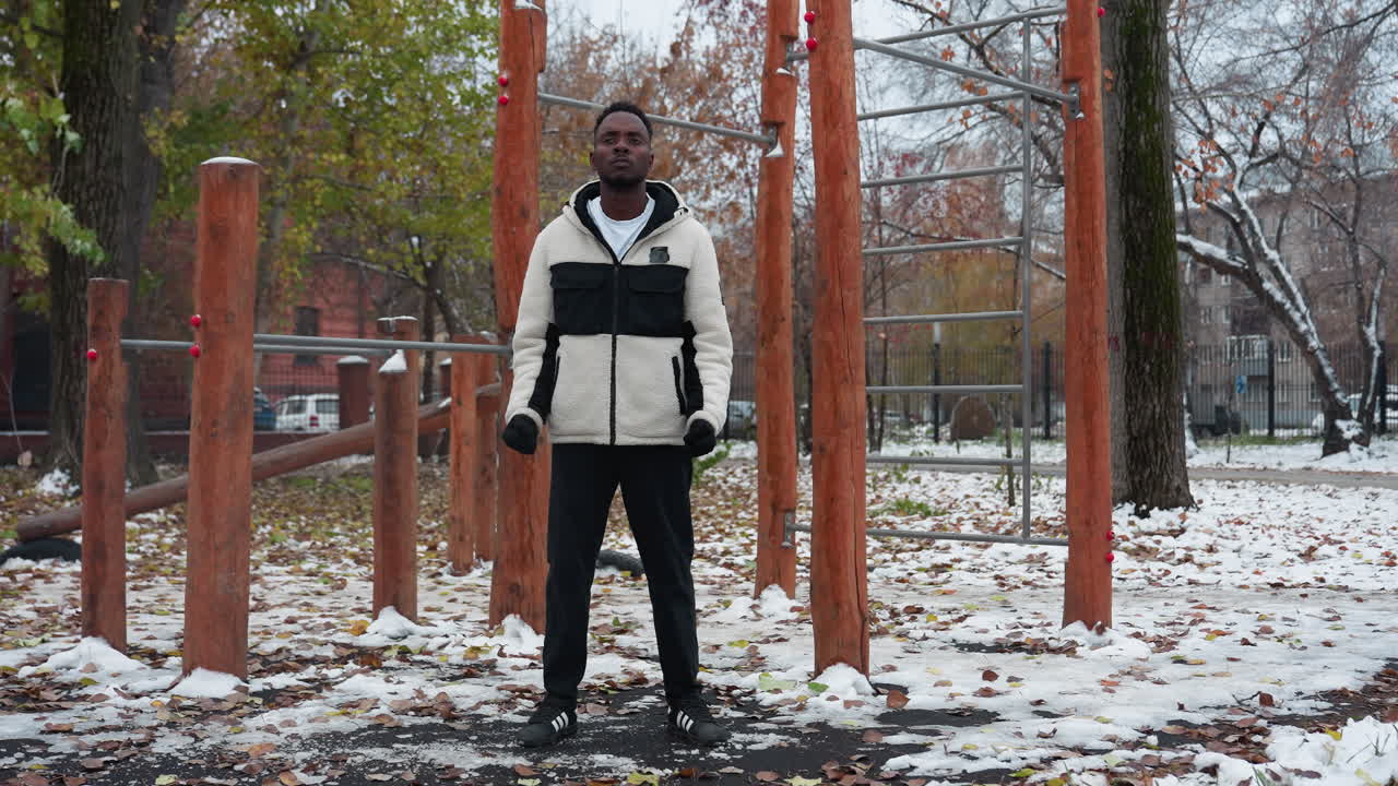 joven adulto con chaqueta de invierno, y pantalones negros realizando torsión de muñeca al aire libre, entorno de parque nevado con equipos de entrenamiento en el fondo, hojas caídas en el suelo, y un coche que pasa lejos