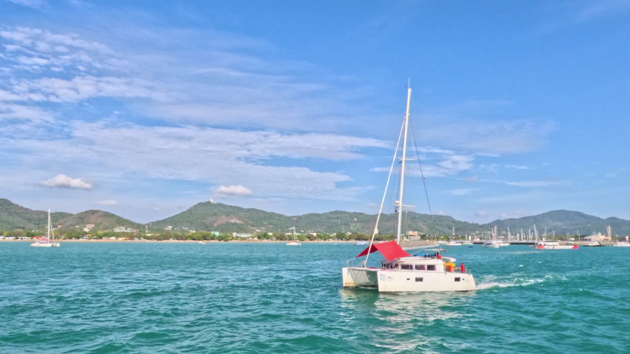 푸켓의 라와이 해변 근처의 푸르키즈색 물을 가로지르는 카타마란. 은 하늘과 멀리 있는 언덕은 평온한 배경을 만니다.