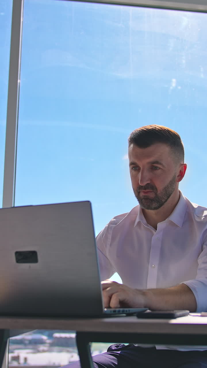 Handsome entrepreneur sitting at table in front of a laptop. Businessman working at his workplace in office on window backdrop. Vertical video