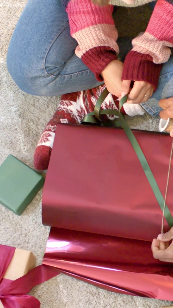 Video verticale: Donna con cappello da Babbo Natale e maglione a casa, che prepara regali con nastro verde e spago.