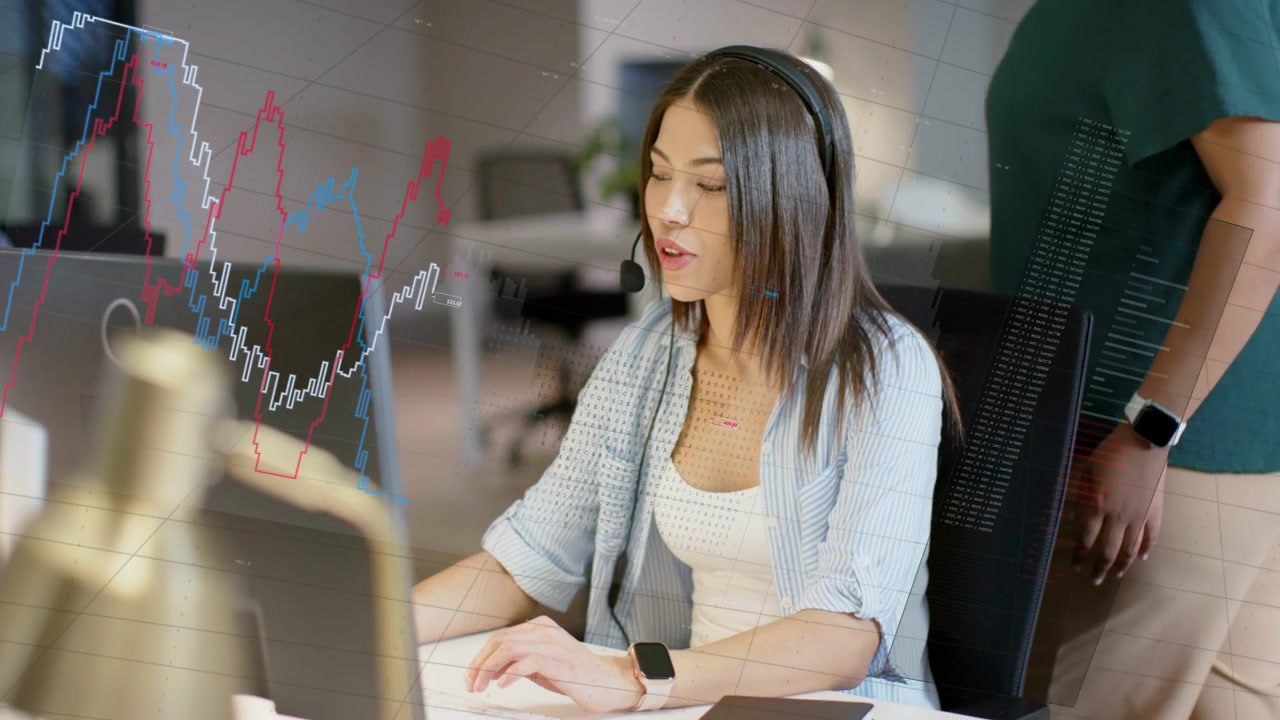 Seated support rep headset on, typing, mouse click showing charts on monitor to help, peer watching