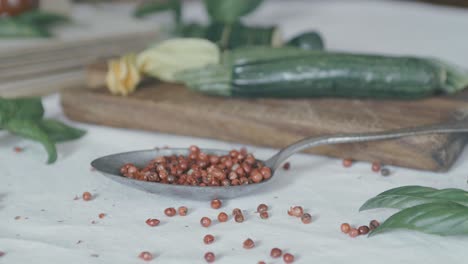 una cuchara de plata llena de pimiento rojo rebota contra la mesa y los granos de pimienta caen por todas partes, también hay calabacines y hojas de albahaca en la escena