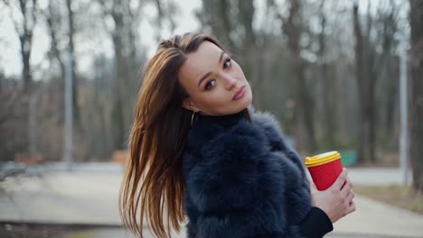 happy dark haired girl in the park. young woman in winter coat walking outdoors and drinking hot beverage in a plastic cup. slow motion.