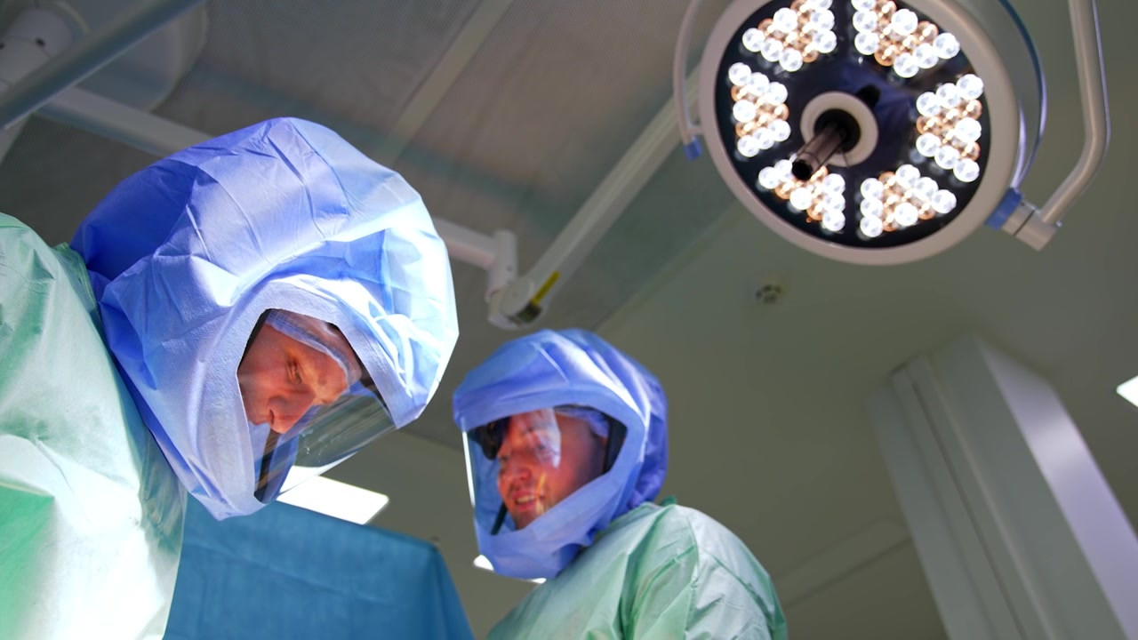 Cirujanos caucásicos con cascos protectores sonríen durante la operación. Vista en ángulo bajo de los médicos trabajando bajo la lámpara redonda.