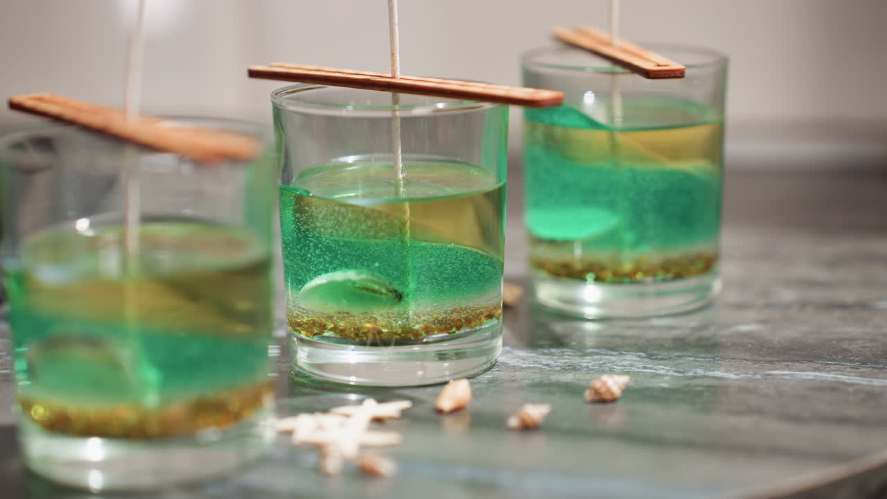 Female creative drops decorative shell-like piece beside transparent glass jars filled with colorful wax and wick setup on kitchen countertop, showcasing artistic candle-making process