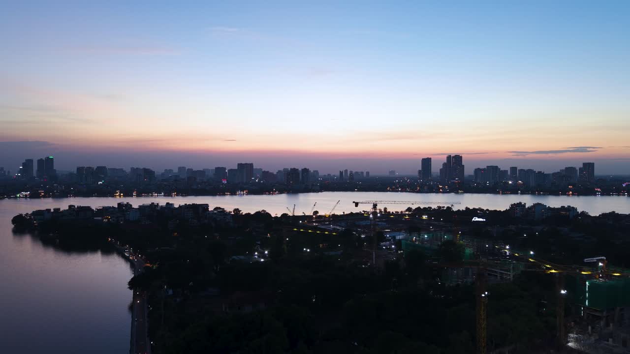 Aerial view of Hanoi's West Lake (Tay Ho) at sunrise. The tranquil water reflects the developing skyline and construction cranes, symbolizing growth and progress