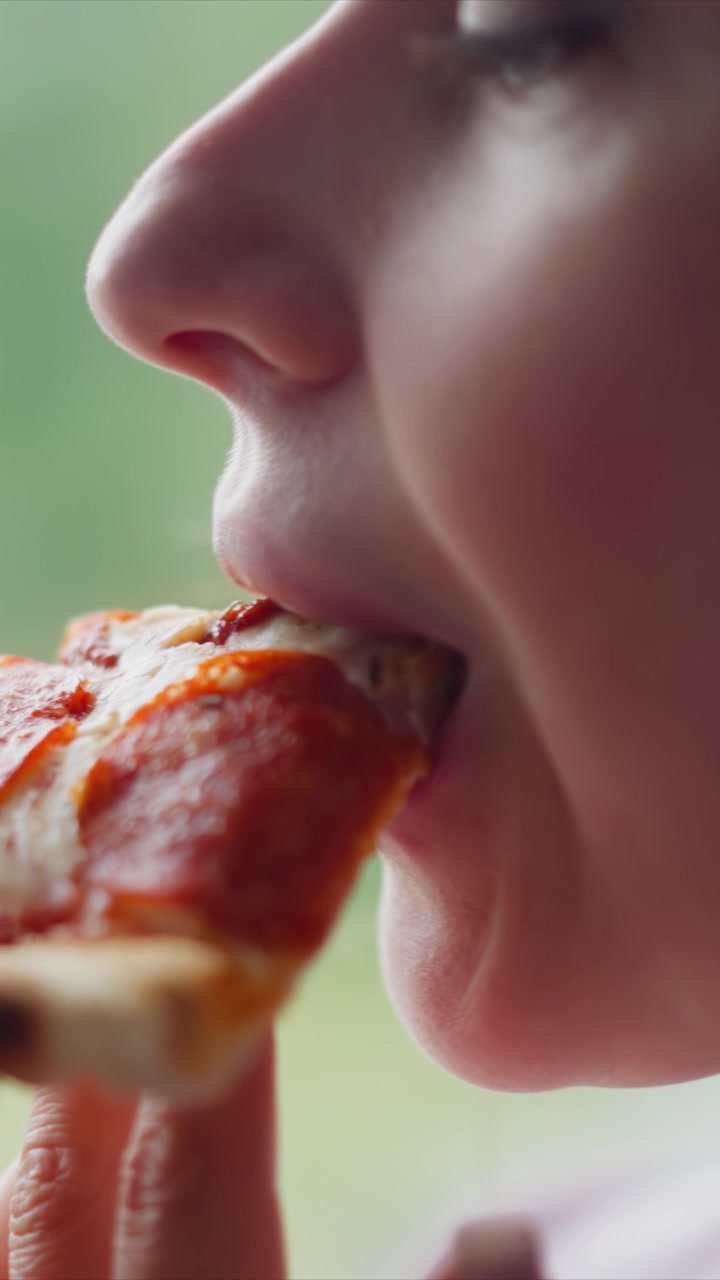 Close up of woman eating pizza. subject holds and takes bites from food with detailed focus on her mouth movements and hand holding slice. Soft lighting creates natural highlights in close