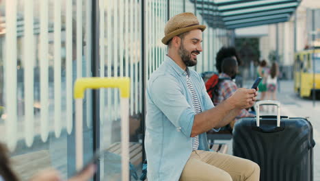 young caucasian handsome man traveller in hat sitting on bench at bus stop and waiting for transport while using smartphone