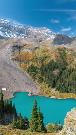 vertical 4k timelapse, turquoise alpine lake under green hills and snow capped mountain peaks on sunny day