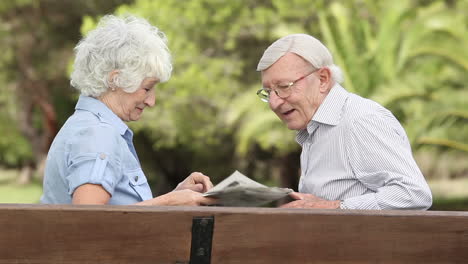 una pareja vieja riendo en un banco con un periódico.