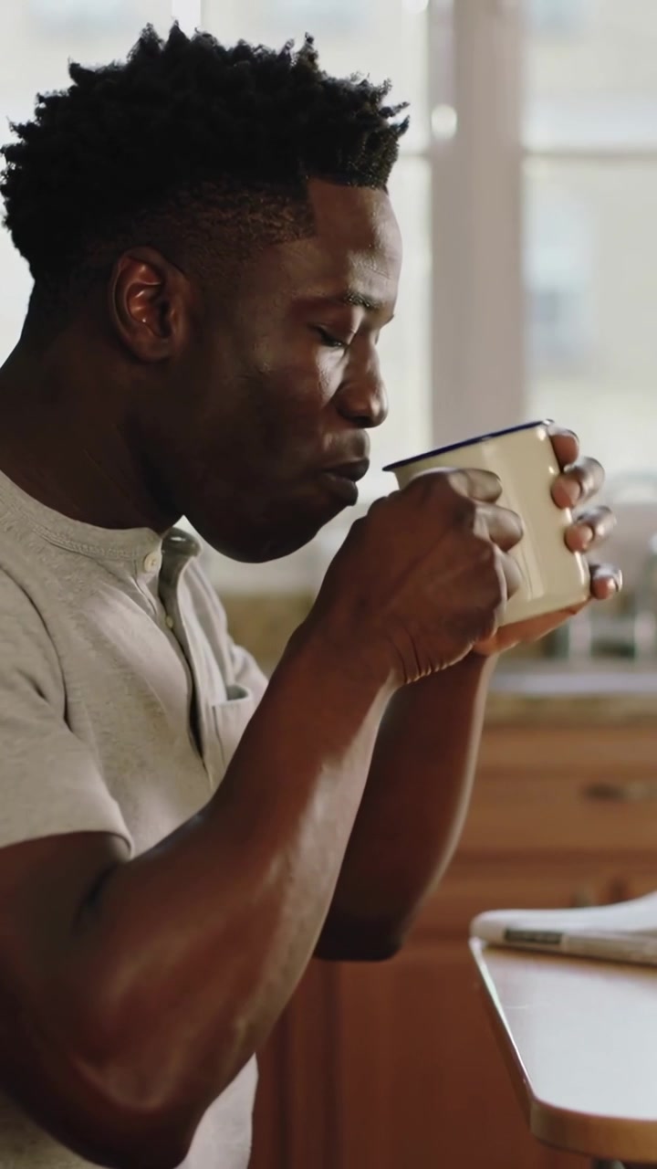 Video verticale: Uomo con maglietta henley che si protende per una tazza smaltata bianca, sorseggiando caffè al tavolo della cucina.