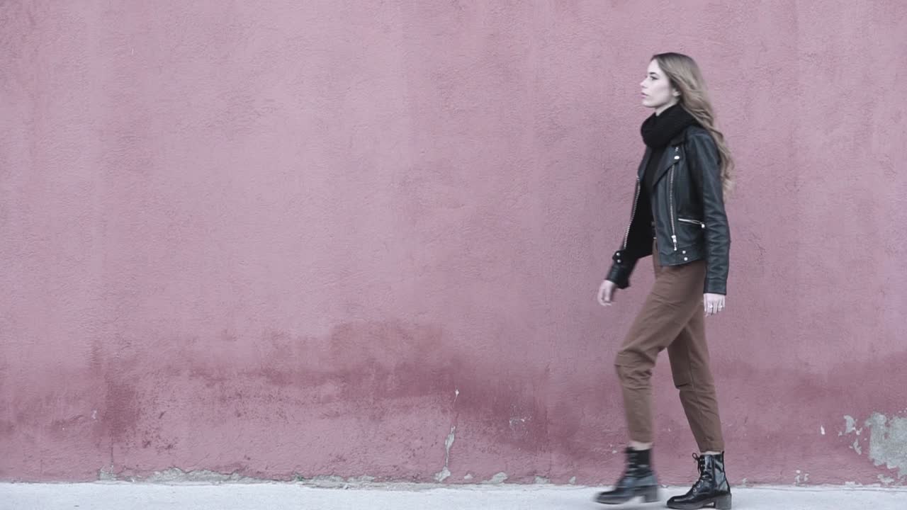 Giovane modella millennial che appare molto sicura di sé mentre cammina in avanti, indossando moderni stivali neri e una giacca di pelle per un look deciso.